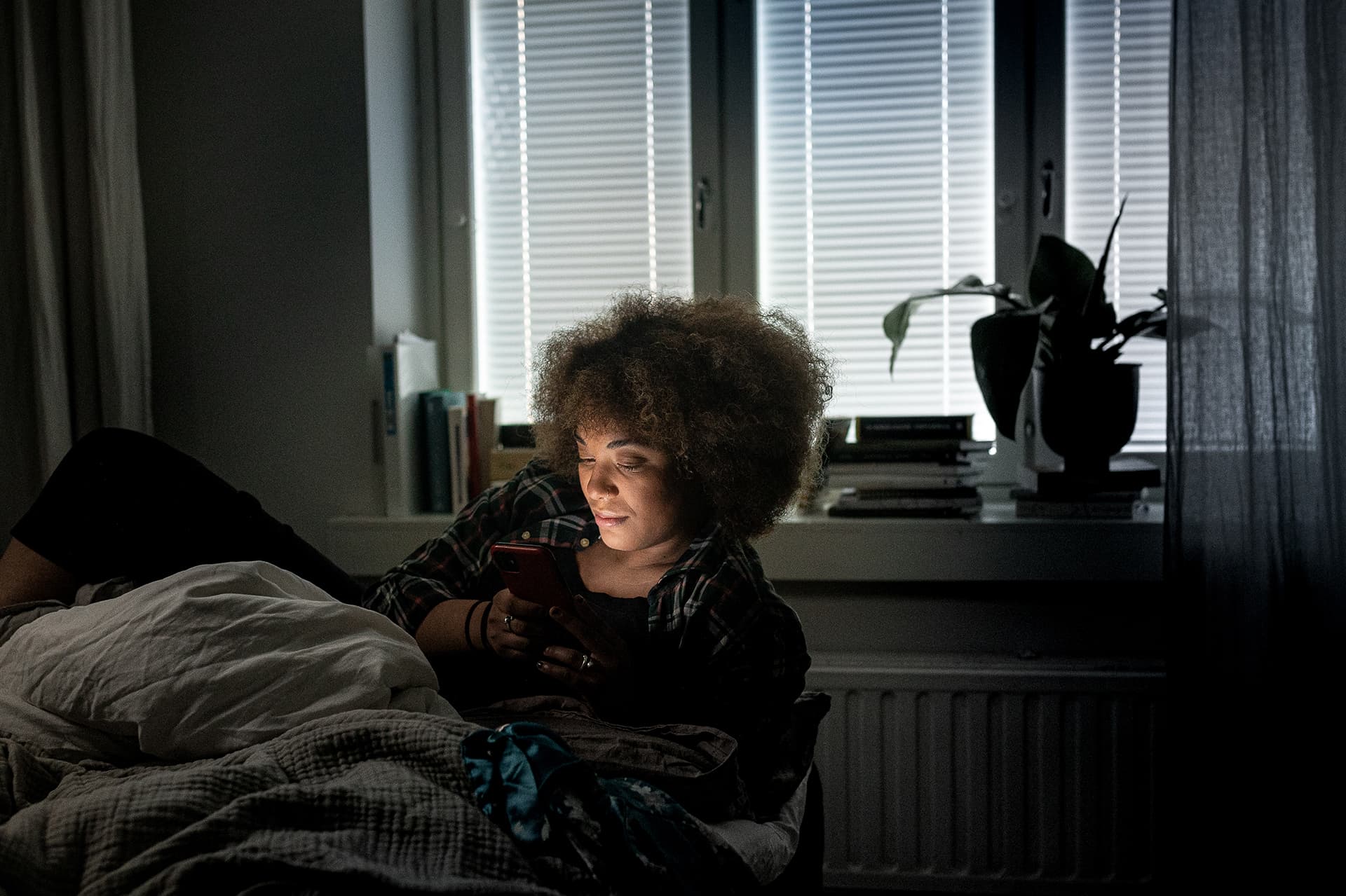 A young woman lying on her side in bed in a dark room and browsing her phone.