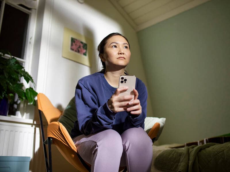 A woman sits thoughtfully in a bedroom armchair with a phone in her hand.