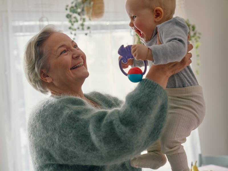 An elderly woman lifts a baby holding toys in its hands.