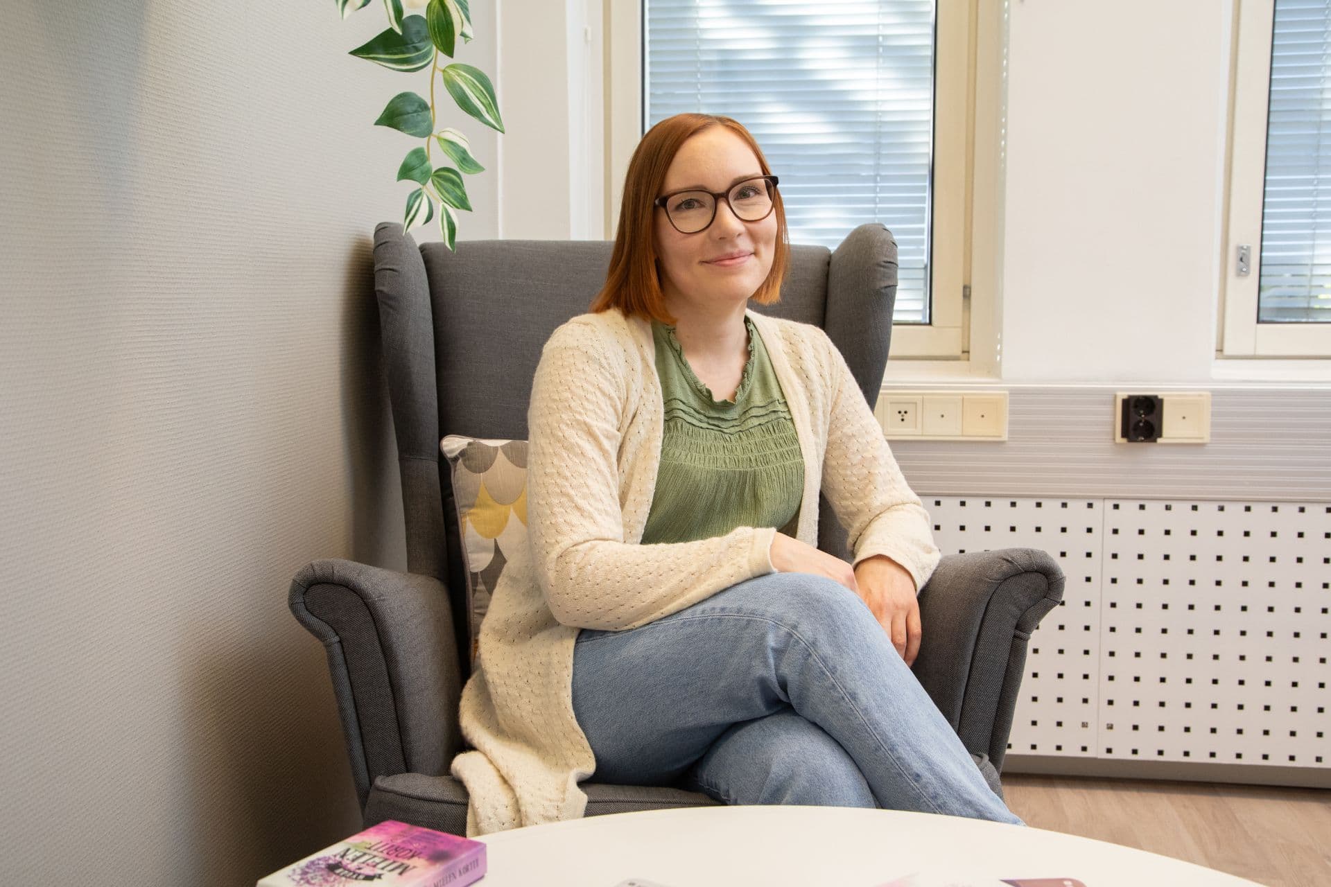 Nainen jolla punertava tukka ja silmälasit istuu hymyillen nojatuolissa rauhallisessa vastaanottohuoneessa ja katsoo kameraan.