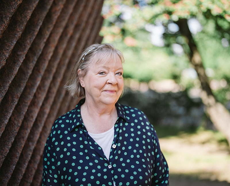 Mehiläinen Kotipalveluiden henkilökohtainen avustaja Leena seisoo puistossa puun varjossa aurinkoisena päivänä.